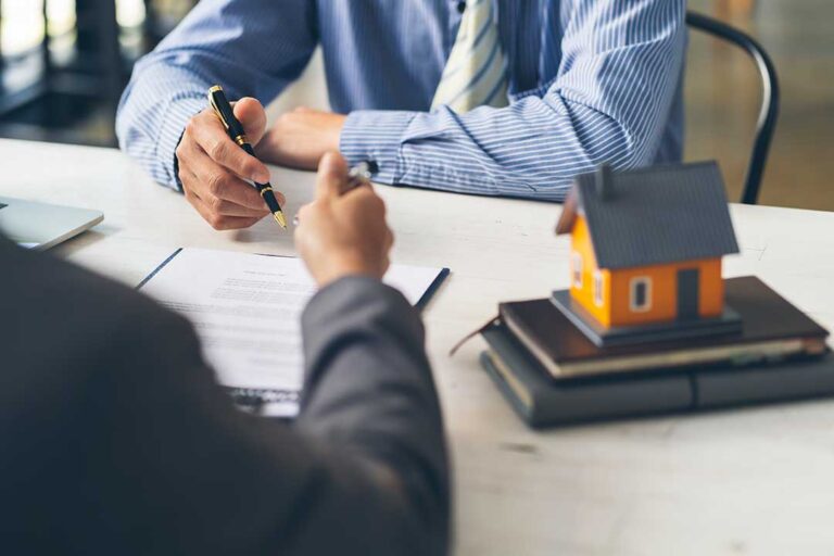 A person reviews mortgage documents with a lender at a kitchen table with a laptop and paper forms.